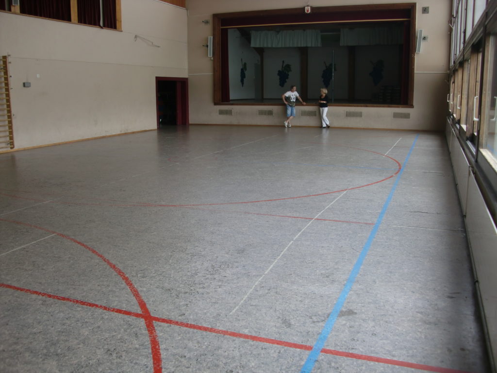 Ancien linoléum gris aux lignes usées dans une salle polyvalente, vue vers la scène