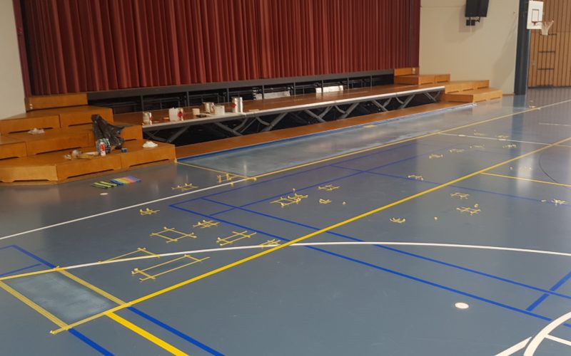 Salle polyvalente pendant la rénovation de son sol, zones délimitées au ruban devant la scène et matériel posé sur les marches