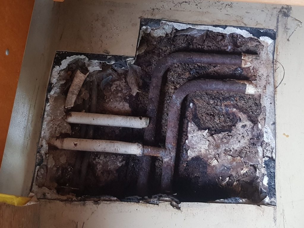 Conduites d'eau rouillées et chape dégradée mises à nu sous le sol d'une salle de rythmique après un dégât d'eau