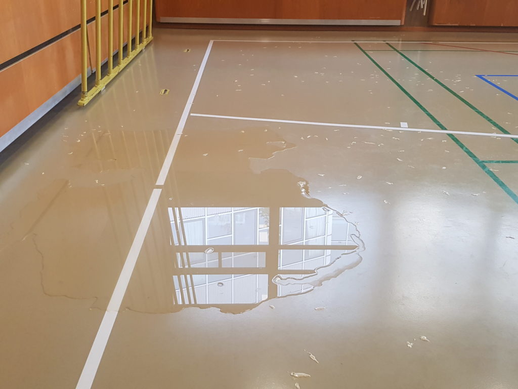 Flaque d'eau de pluie infiltrée par la toiture sur un sol sportif dont les fissures ont été traitées la veille