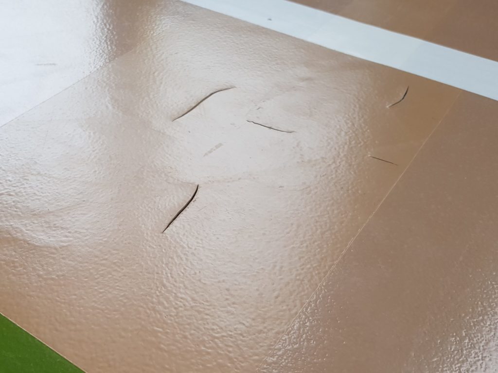 Fissures ouvertes dans le revêtement d'un sol sportif sans joint, près d'une ligne de jeu verte