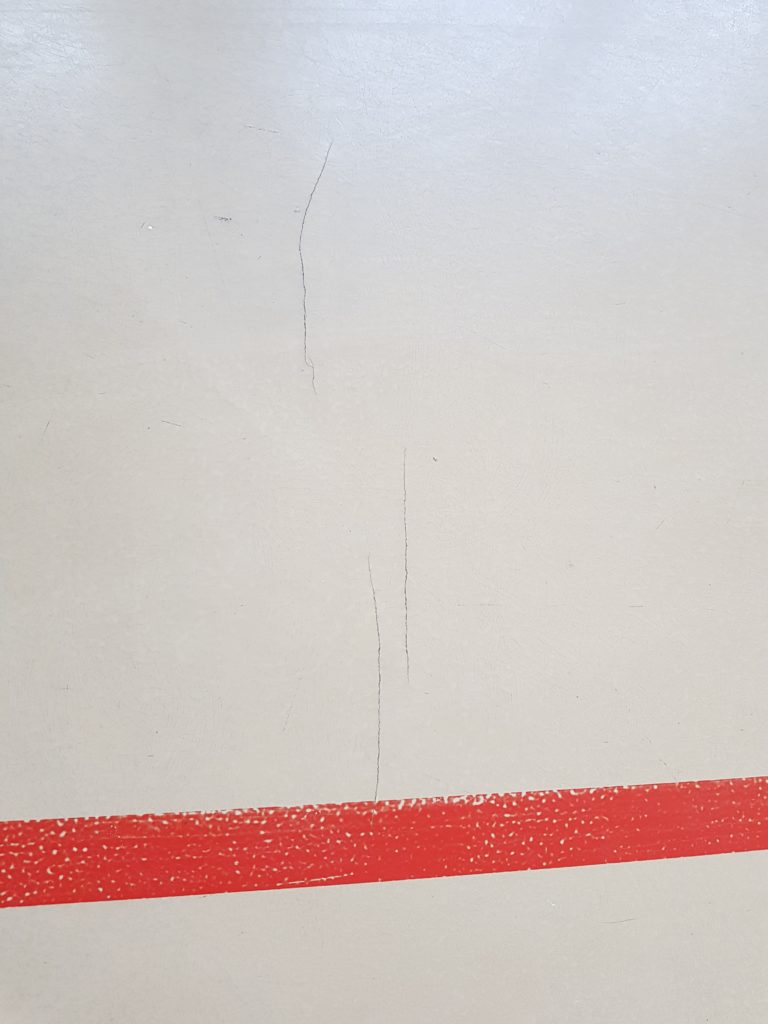 Fissures ouvertes dans le revêtement en résine d'un sol sportif, près d'une ligne de jeu rouge