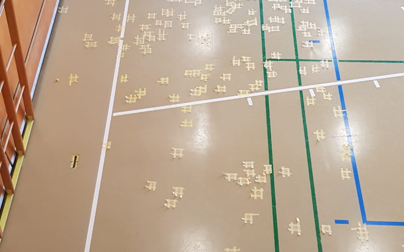 Vue en hauteur d'une salle de gymnastique dont les nombreuses fissures du sol sont traitées en urgence, chaque réparation délimitée par du ruban de masquage