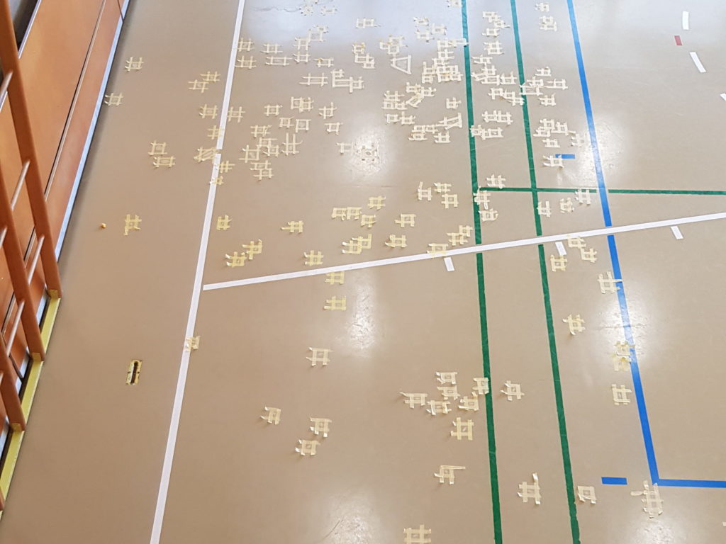 Vue en hauteur d'une salle de gymnastique dont les nombreuses fissures du sol sont traitées en urgence, chaque réparation délimitée par du ruban de masquage