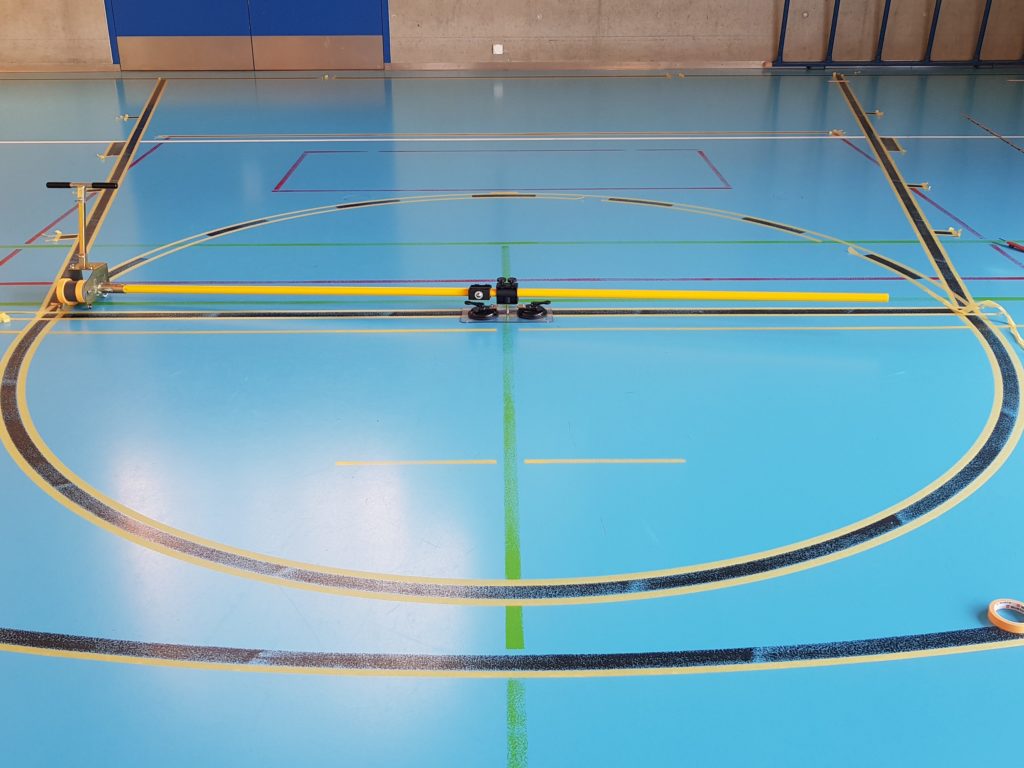 Marquage en cours autour du rond central d'un sol sportif, lignes masquées au ruban et traceur de cercle fixé au centre
