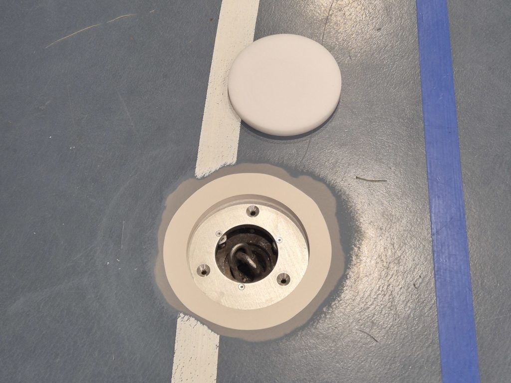 Nouvelle embase métallique vissée dans un sol sportif, couvercle de douille d'origine posé à côté avant sa réintégration