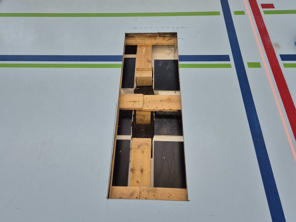 Ouverture rectangulaire dans un sol sportif laissant apparaître les lambourdes et l'isolation de la sous-construction