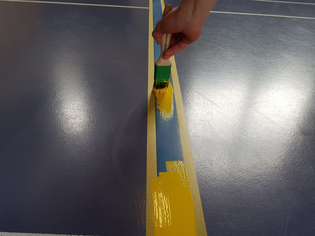 Nouvelle ligne de jeu peinte en jaune au pinceau entre deux rubans, par-dessus l'ancienne ligne bleue