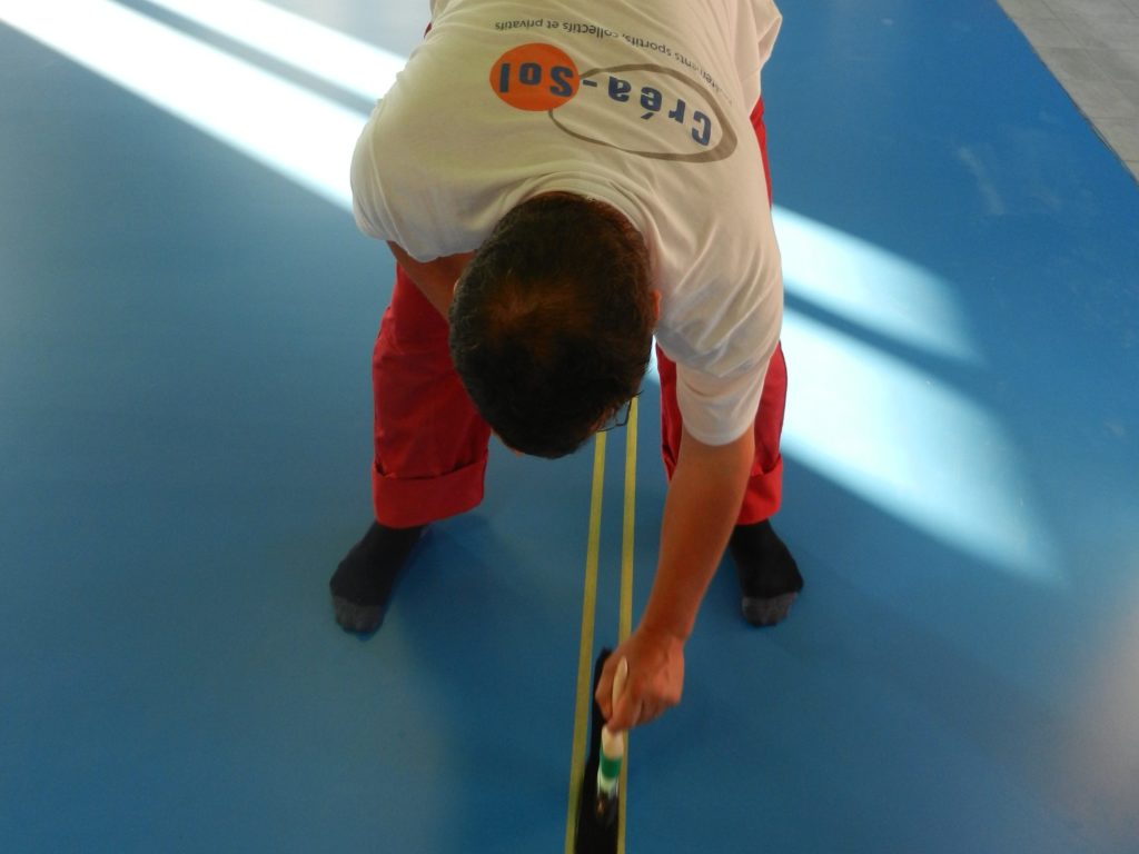 Peinture d'une ligne de jeu noire au pinceau entre deux bandes de scotch sur un sol sportif bleu