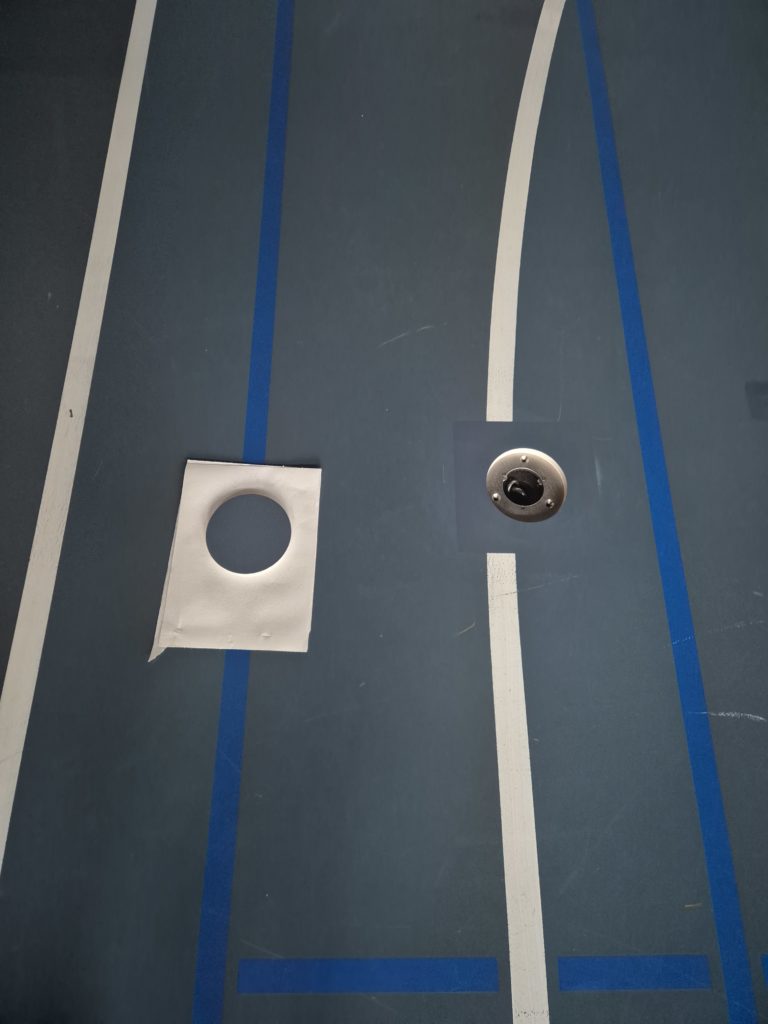 Zone repeinte autour d'une douille réparée sur un sol sportif, embase visible et gabarit de peinture posé à côté