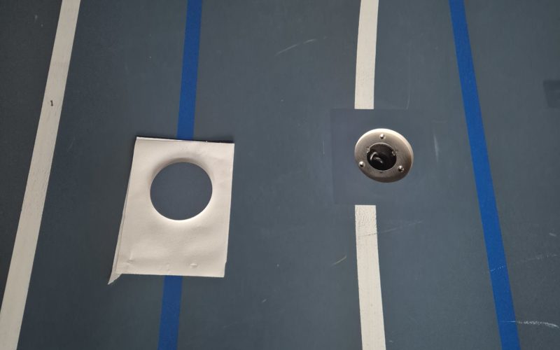 Zone repeinte autour d'une douille réparée sur un sol sportif, embase visible et gabarit de peinture posé à côté