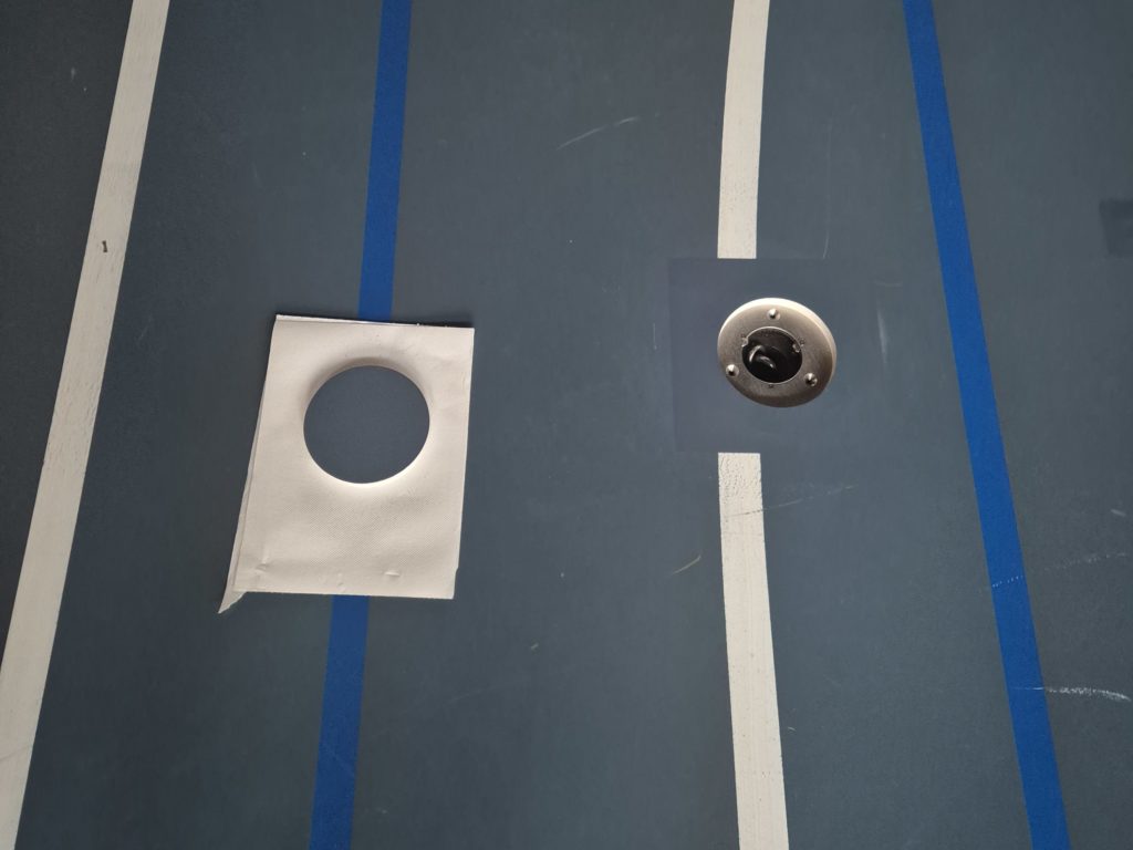 Zone repeinte autour d'une douille réparée sur un sol sportif, embase visible et gabarit de peinture posé à côté