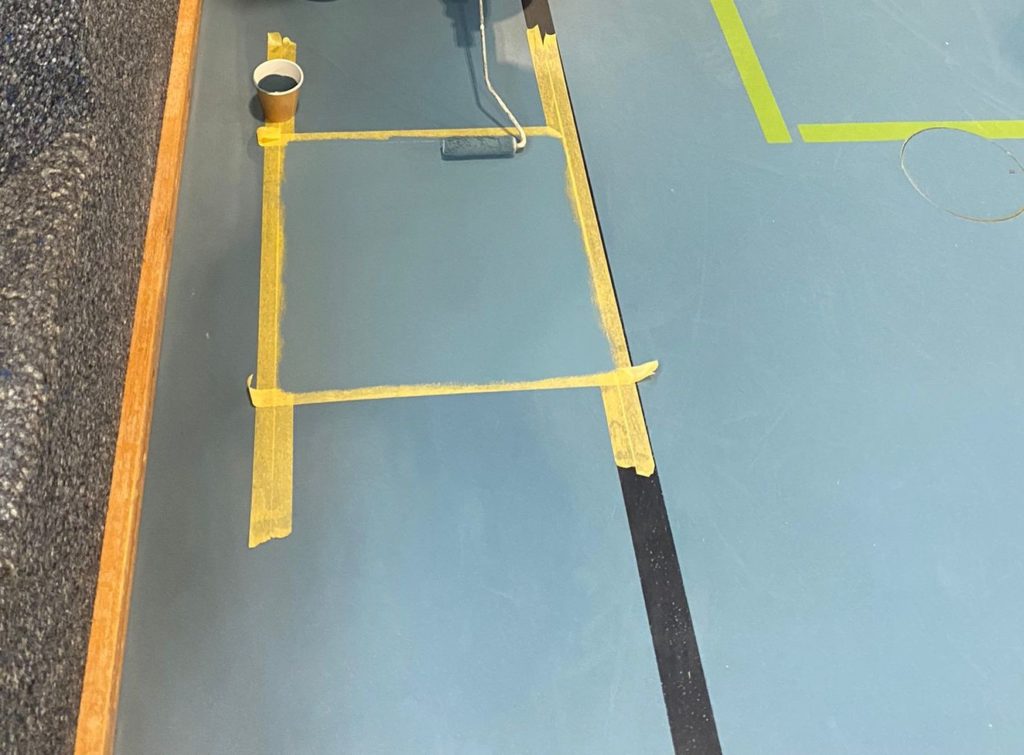 Peinture appliquée au rouleau sur la zone réparée d'un sol sportif, délimitée par du ruban de masquage