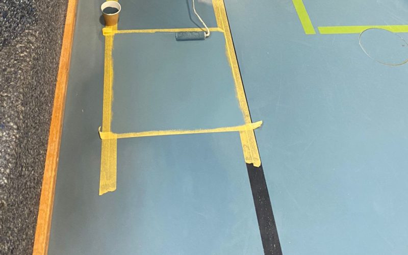 Peinture appliquée au rouleau sur la zone réparée d'un sol sportif, délimitée par du ruban de masquage