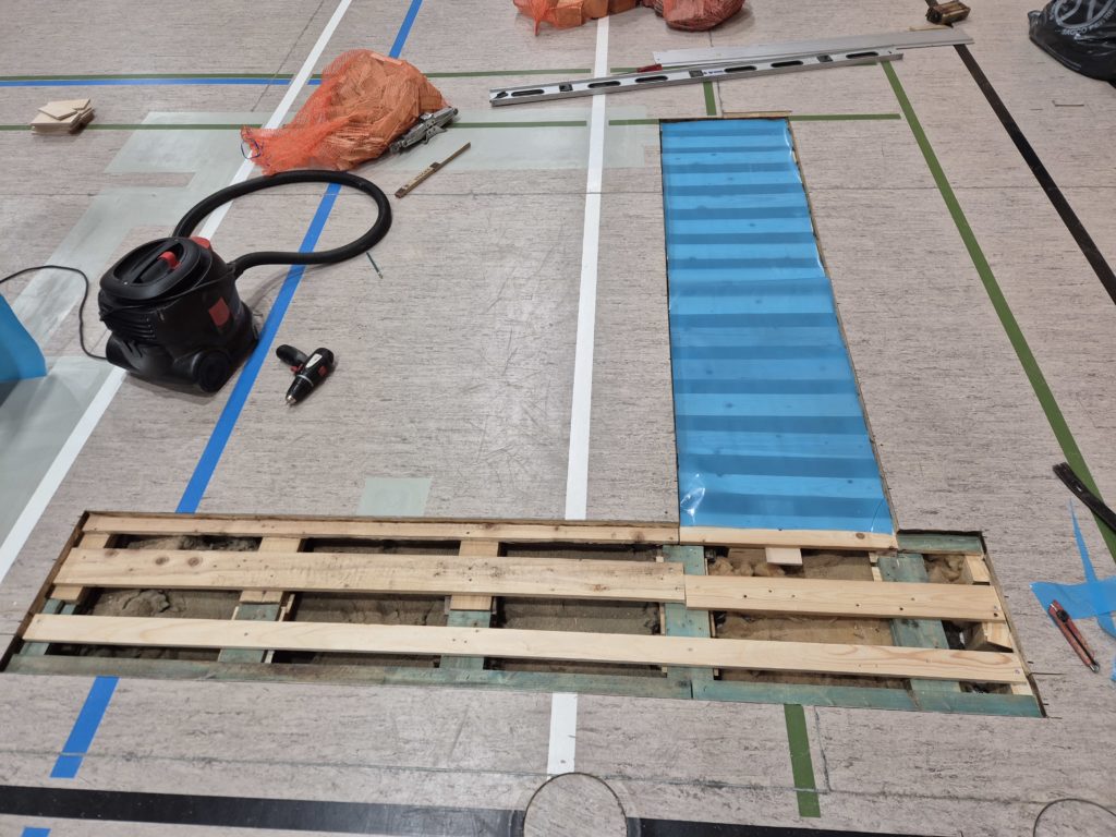 Nouvelle sous-construction en bois raccordée aux lambourdes d'origine sous un sol sportif en linoléum, zone voisine refermée par des panneaux