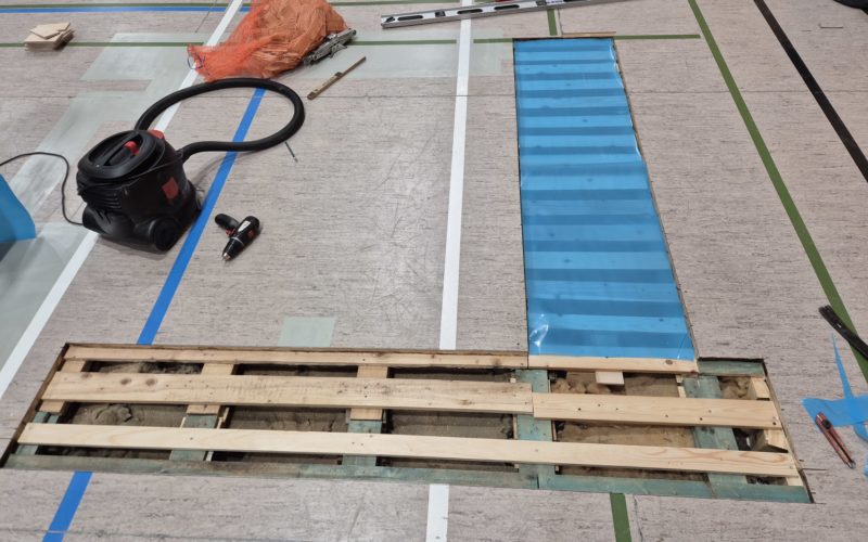 Nouvelle sous-construction en bois raccordée aux lambourdes d'origine sous un sol sportif en linoléum, zone voisine refermée par des panneaux