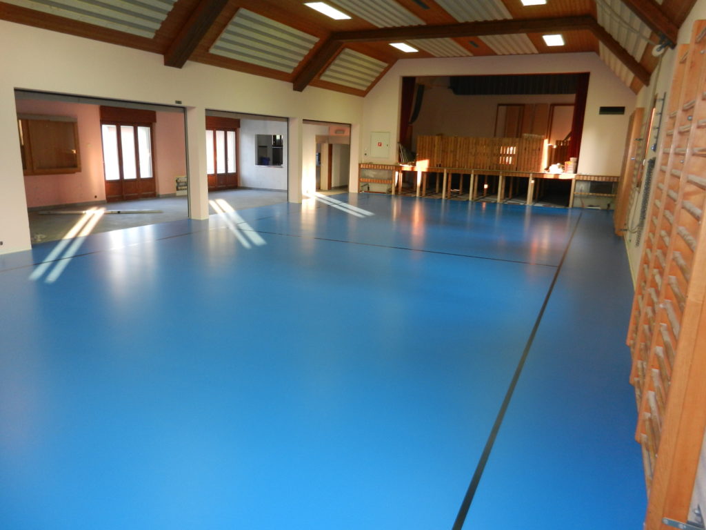 Salle polyvalente rénovée avec un sol sportif bleu, vue vers la scène