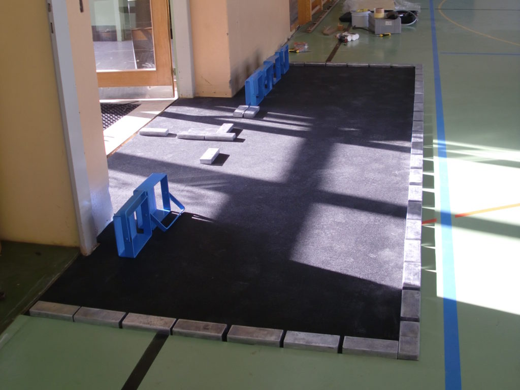 Nouvelle sous-couche en caoutchouc collée dans la zone reconstruite d'un sol sportif, lestée par des poids pendant le séchage