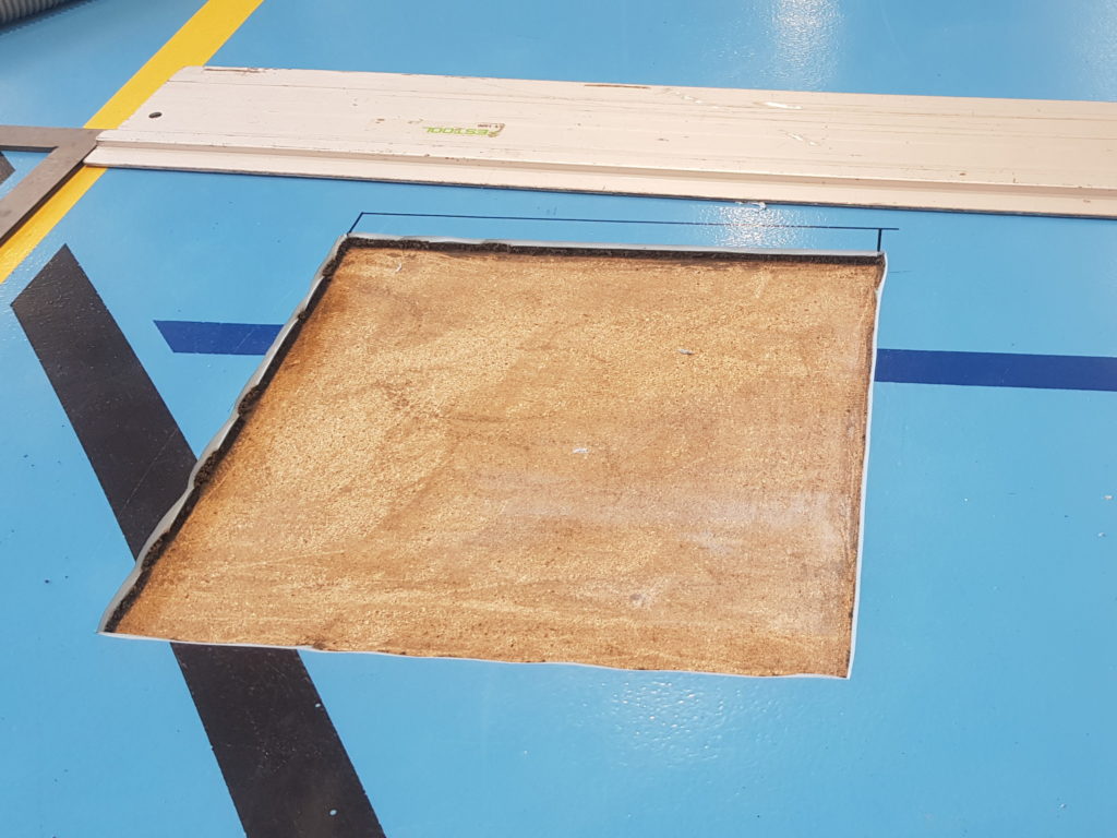 Zone cloquée découpée dans un sol sportif, revêtement retiré et support apparent, rail de guidage posé à côté
