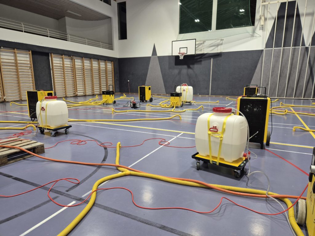 Assèchement d'un sol sportif à souplesse combinée après une fuite d'eau : déshumidificateurs et gaines raccordées aux percements dans une salle polyvalente