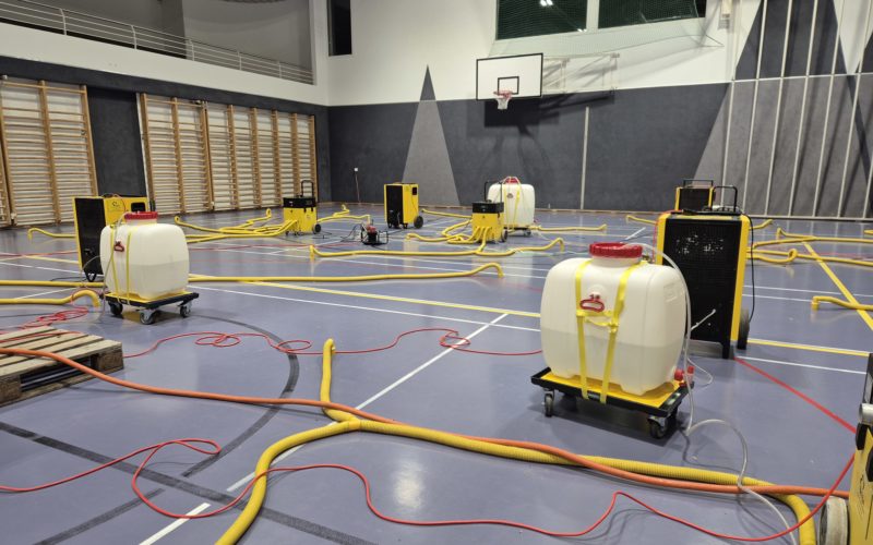 Assèchement d'un sol sportif à souplesse combinée après une fuite d'eau : déshumidificateurs et gaines raccordées aux percements dans une salle polyvalente