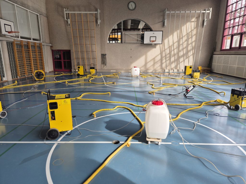 Assèchement d'un sol sportif après un dégât d'eau dû à un orage : déshumidificateurs et gaines raccordées aux percements dans une salle de gymnastique