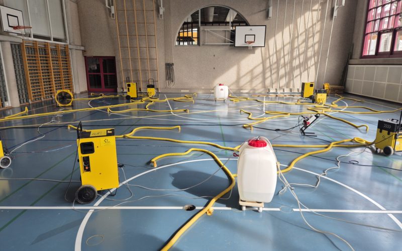 Assèchement d'un sol sportif après un dégât d'eau dû à un orage : déshumidificateurs et gaines raccordées aux percements dans une salle de gymnastique