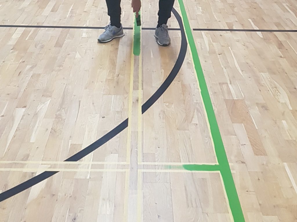 Peinture d'une ligne de jeu verte au pinceau entre deux bandes de scotch, sur le parquet vitrifié.