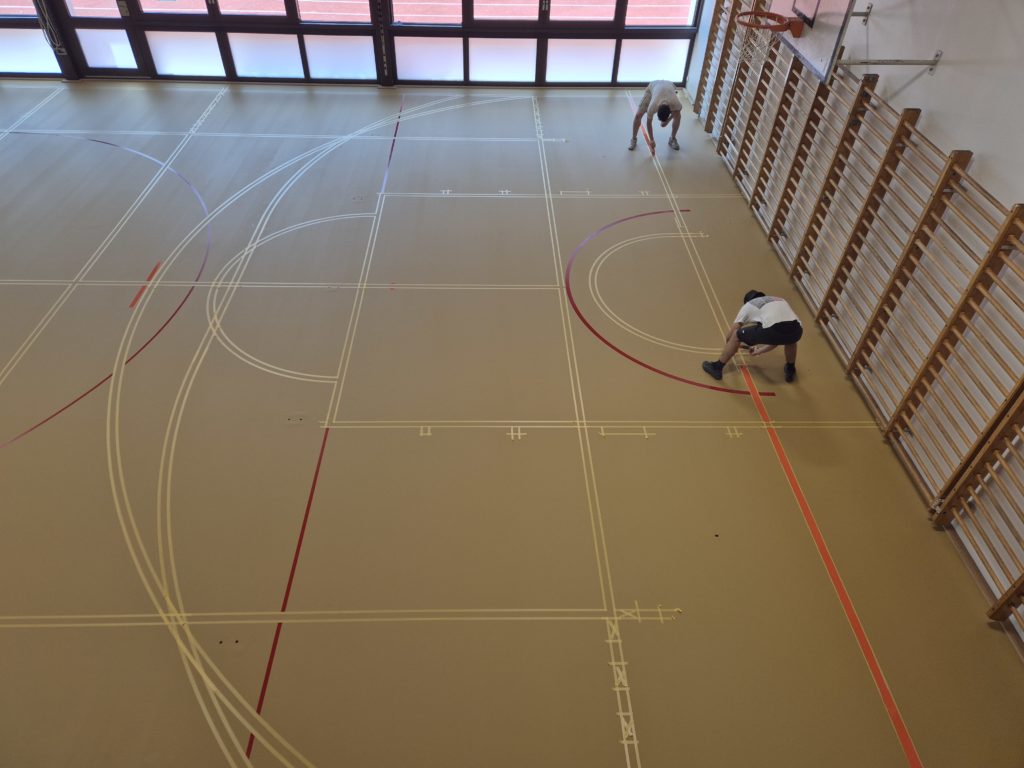 Peinture des premières lignes de jeu entre les bandes de scotch sur le sol sportif rénové.