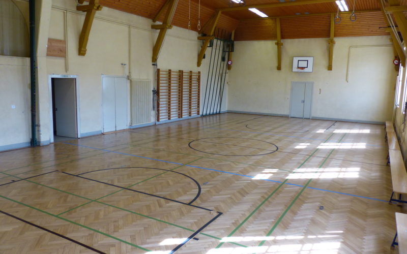 Salle de gymnastique au parquet en chêne en bâtons rompus rénové, avec un marquage pour plusieurs sports.