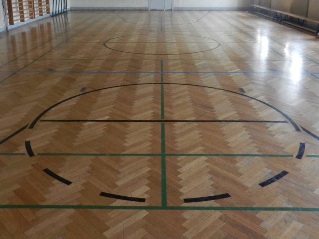 Salle de gymnastique au parquet en chêne en bâtons rompus avant rénovation, bois foncé et lignes de jeu usées.