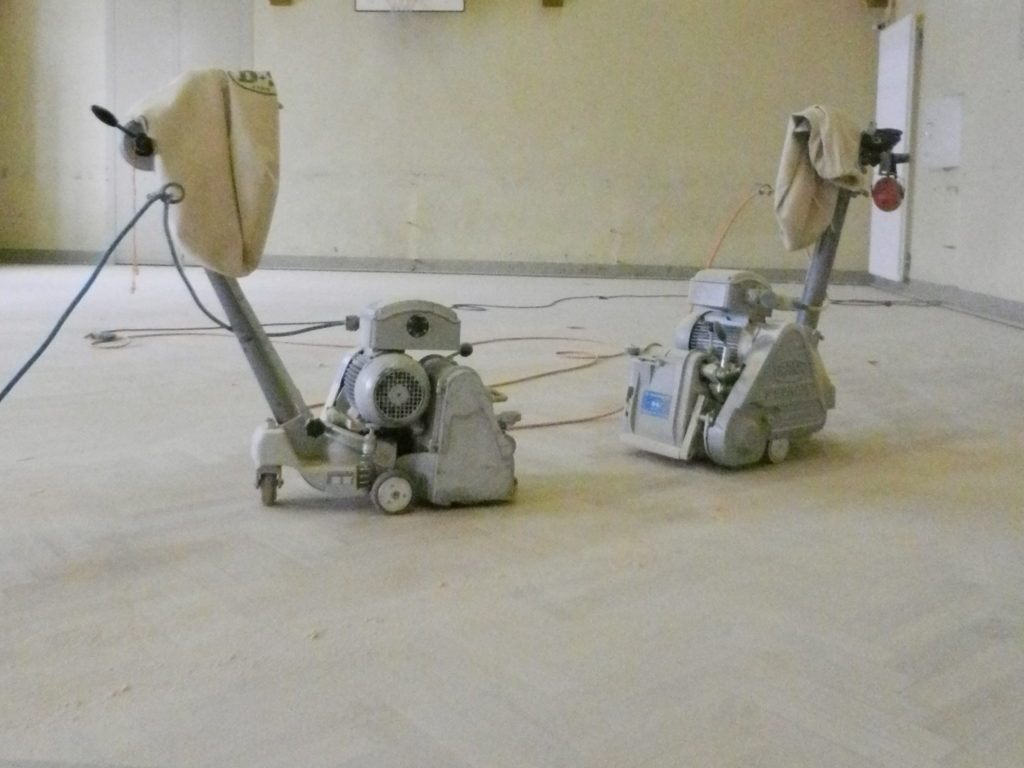 Deux ponceuses à bande sur le parquet poncé jusqu'au bois clair, anciennes lignes retirées.