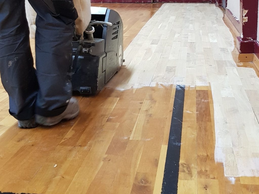Ponçage du parquet à la ponceuse à bande : le bois clair réapparaît à côté de l'ancienne vitrification et d'une ligne usée.