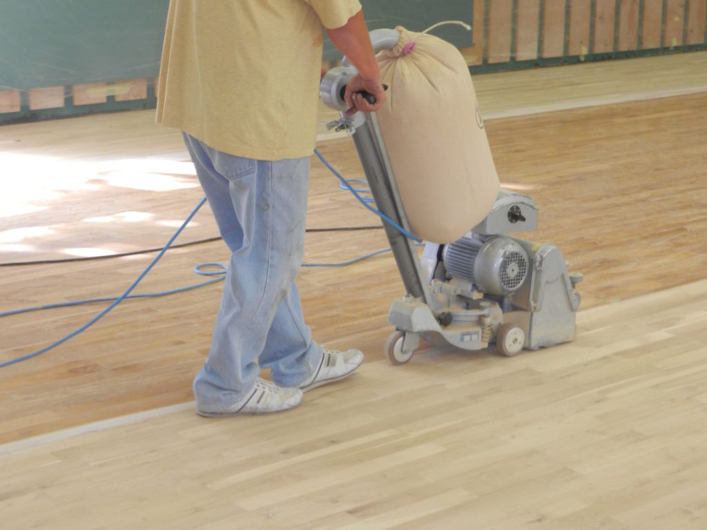 Ponçage du parquet sportif à la ponceuse à bande jusqu'au bois clair, murs protégés par un film plastique.