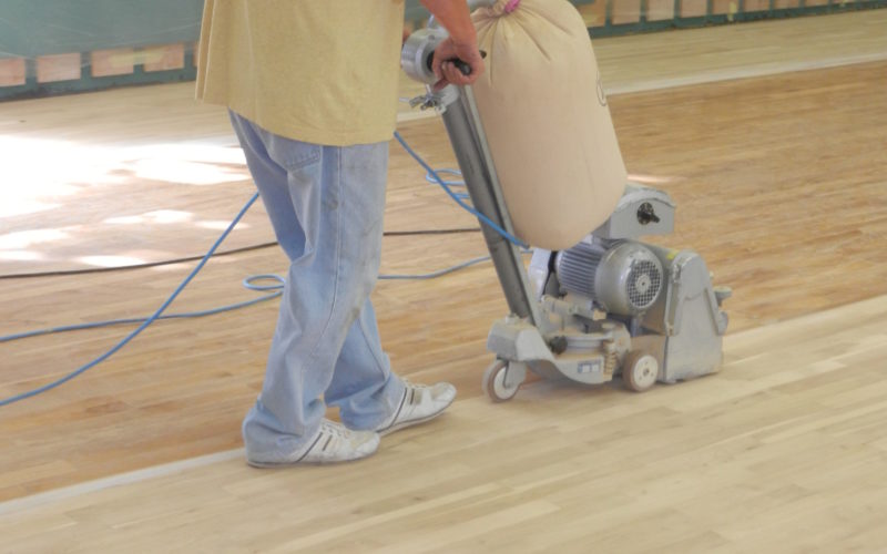 Ponçage du parquet sportif à la ponceuse à bande jusqu'au bois clair, murs protégés par un film plastique.