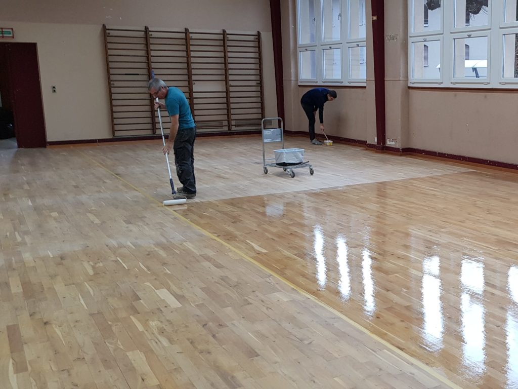 Application de la vitrification au rouleau sur le parquet sportif poncé, bordures reprises au pinceau.